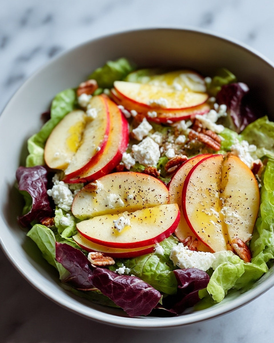 A salad in a white bowl resting on a white marbled surface, made up of three main layers: the bottom layer has fresh green and red lettuce leaves with crisp texture, the middle layer shows thin slices of red apple with yellow inside arranged in a circular pattern, and the top layer is scattered with white crumbled cheese and small brown nuts. There is a light drizzle of olive oil and a sprinkling of black pepper visible on the apple slices and cheese, giving the salad a fresh and colorful look. photo taken with an iphone --ar 4:5 --v 7