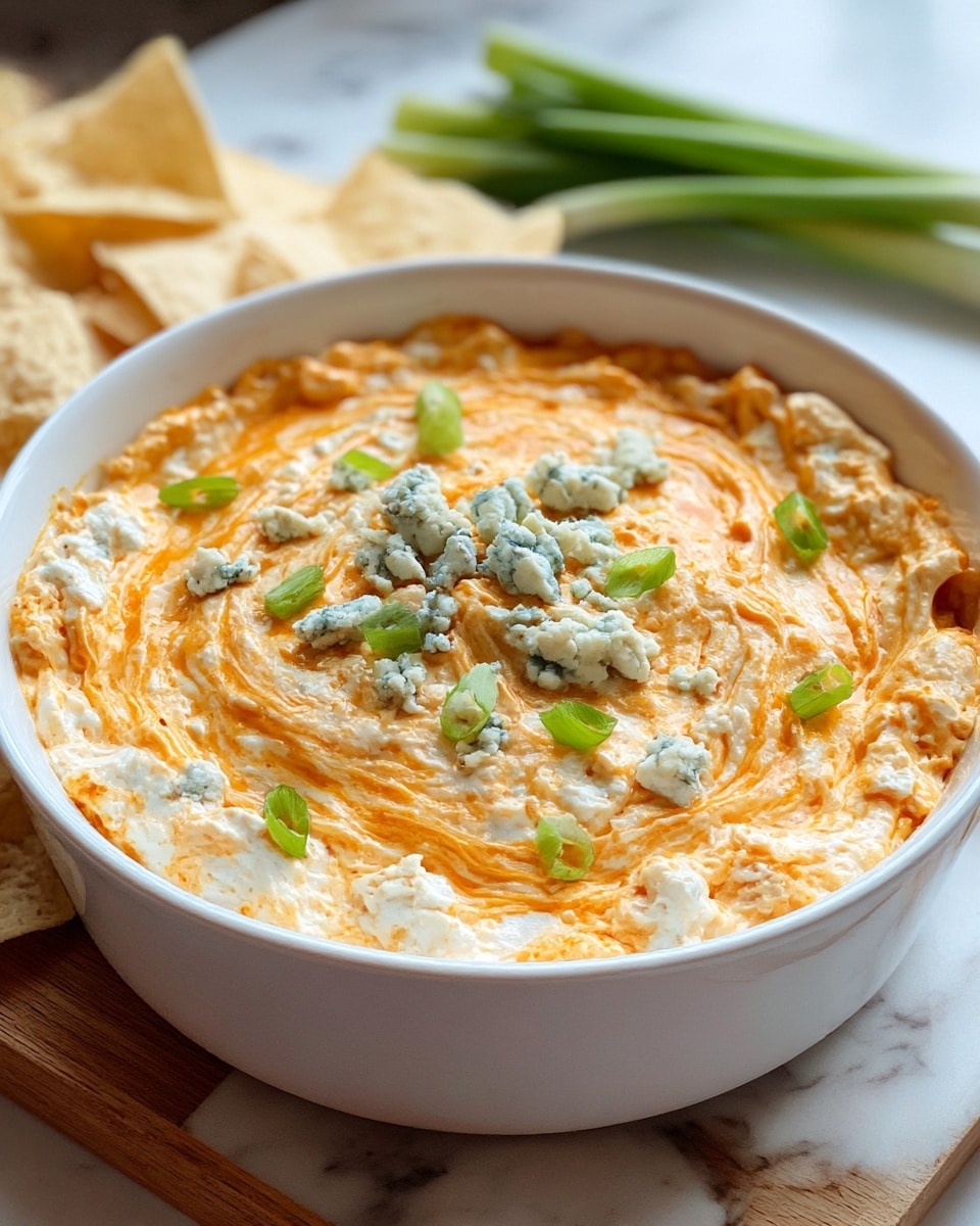 A close-up view of a white round dish filled with a creamy, orange and white layered dip with a slightly rough texture, topped in the center with small crumbles of blue cheese and scattered green onion pieces. The creamy orange layer appears swirled and glossy, sitting on a base mixture that has a chunkier texture with white and light orange colors. In the background are some light beige tortilla chips and fresh green onions resting on a white marbled surface. The dish is placed on a wooden board. Photo taken with an iphone --ar 4:5 --v 7