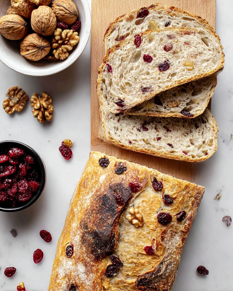 The image shows a rustic loaf of bread on a white marbled surface, with a golden-brown crust featuring darker toasted spots and visible pieces of dried cranberries and walnuts embedded in the crust. Two slices of the bread lie above the loaf, revealing an airy, light beige crumb inside speckled with red dried cranberries and light brown walnut chunks distributed evenly. To the left, there is a white wooden bowl filled with walnut halves and a small black cup filled with dried cranberries, with some nuts and cranberries scattered around on the surface. photo taken with an iphone --ar 4:5 --v 7