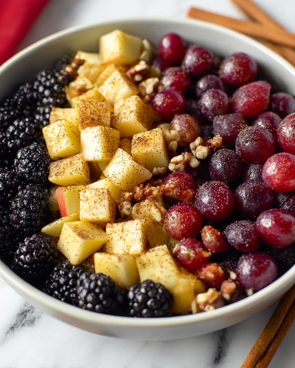 A dark gray bowl is filled with a fresh fruit salad showing at least three layers mixed together: glossy deep red grapes, plump blackberries with a textured surface, and light yellow apple cubes with red skin edges, all sprinkled with small brown pecan pieces and a dusting of cinnamon powder that gives a warm tone to the fruit. The bowl sits on a white marbled surface, accompanied by two cinnamon sticks at the bottom left and a metal whisk and red patterned cloth slightly blurred in the background. Photo taken with an iphone --ar 4:5 --v 7