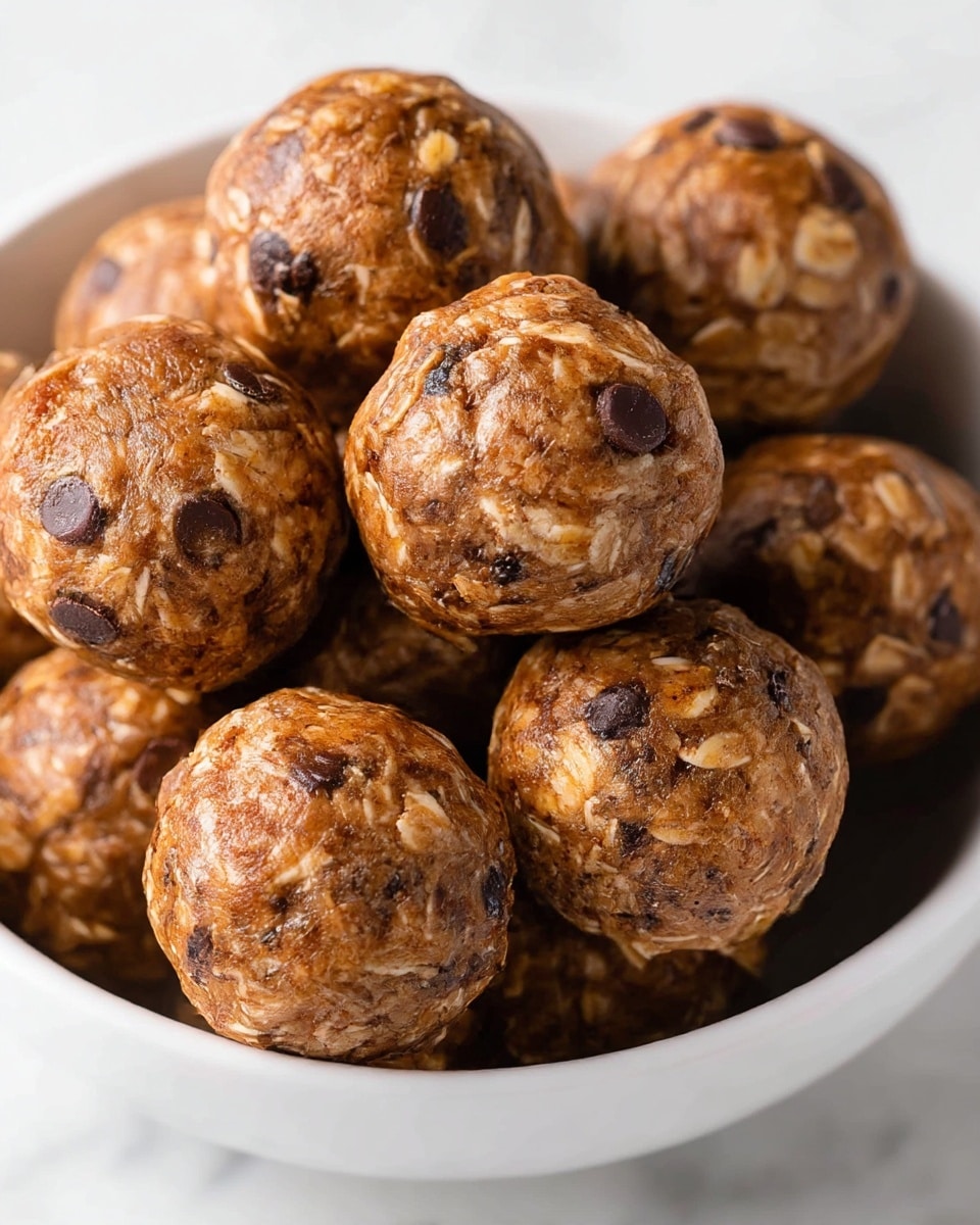 The image shows a close-up of a bunch of round energy balls clustered together inside a white bowl. Each ball has a textured surface with visible oats and small dark chocolate chips embedded throughout, giving them a mix of light brown and dark brown spots. The balls appear moist and dense, piled in layers so they slightly overlap. The bowl sits on a white marbled surface which contrasts with the warm tones of the energy balls. photo taken with an iphone --ar 4:5 --v 7