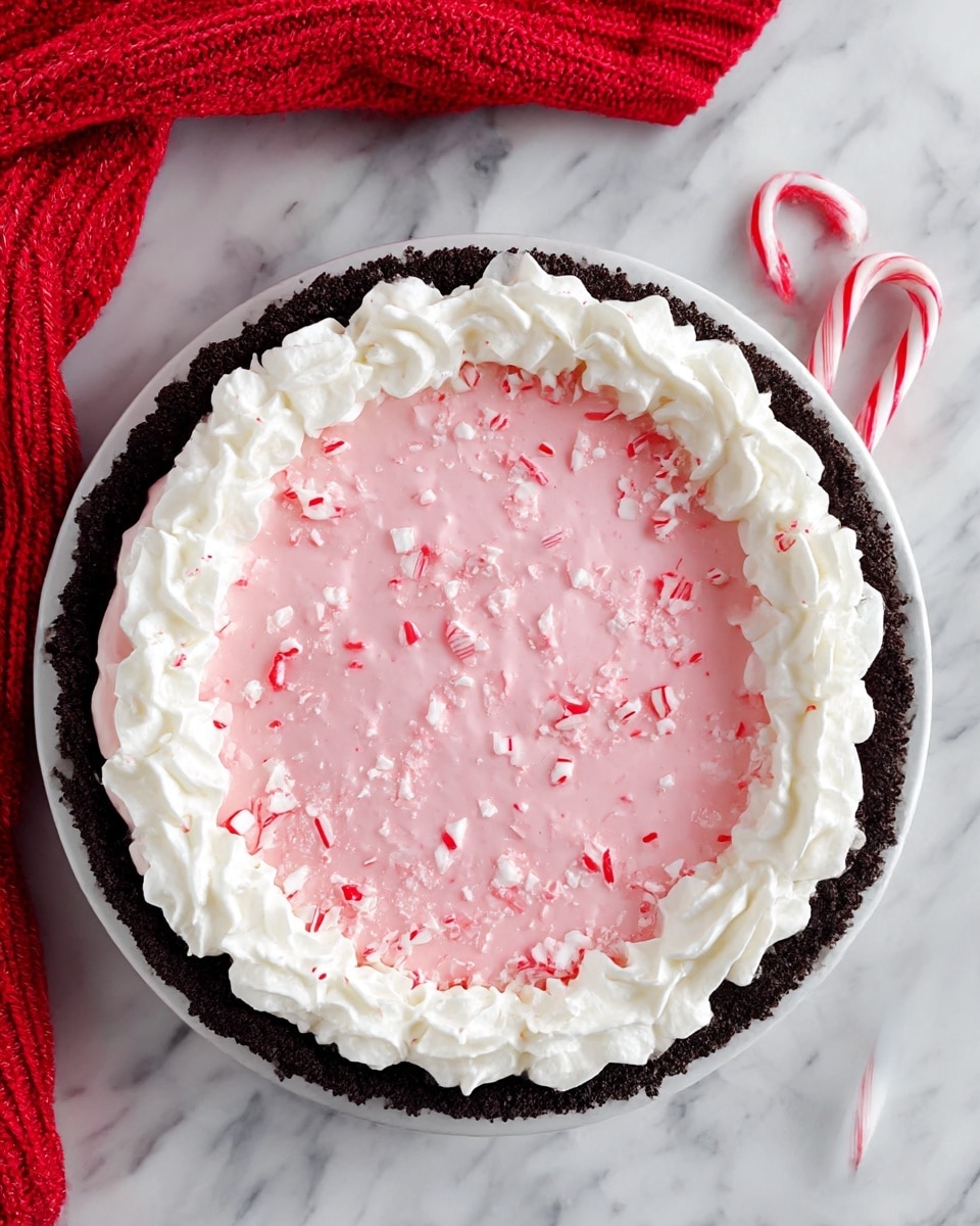 The image shows a round pie placed on a white marbled surface. The pie has three distinct layers: the bottom layer is a dark brown crumbly crust forming the base and sides, the middle layer is a smooth, creamy pink filling covering the entire surface inside the crust, and the top layer features a ring of white whipped cream piped evenly around the edge. Scattered across the pink filling are small, crushed bits of red and white candy, adding texture and color contrast. Next to the pie are two red and white striped candy canes and a piece of red knitted fabric. photo taken with an iphone --ar 4:5 --v 7