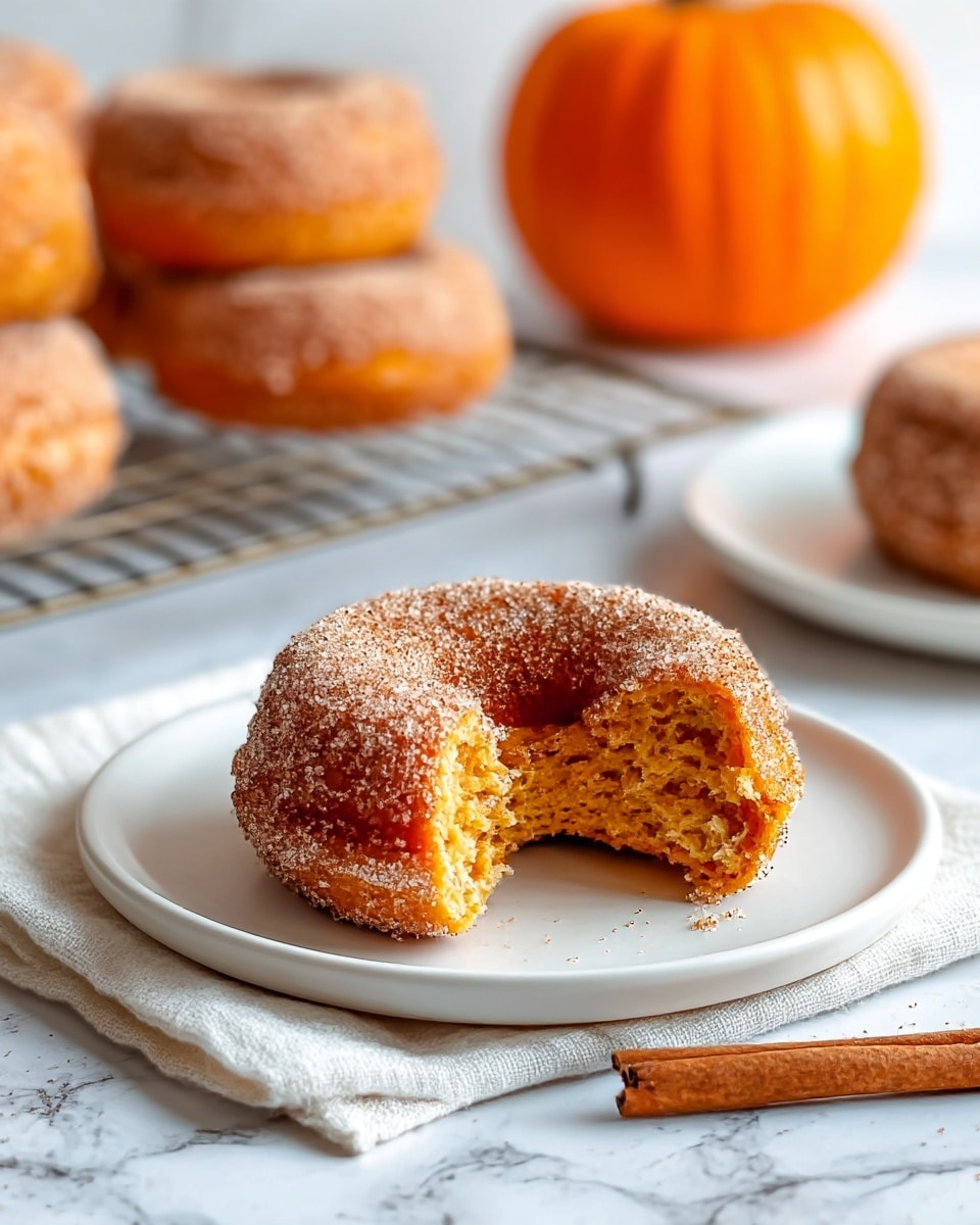 A close-up of a single donut with a missing bite, showing an orange-brown, soft textured inside layer, covered fully with a rough cinnamon sugar coating sprinkled on the outside layer. The donut sits on a white plate resting on a folded white cloth. In the background, there are more similar donuts on a wire rack and a white plate, with a bright orange pumpkin slightly blurred behind them. A single cinnamon stick lies near the plate on a white marbled surface. The scene is well-lit with natural light. photo taken with an iphone --ar 4:5 --v 7