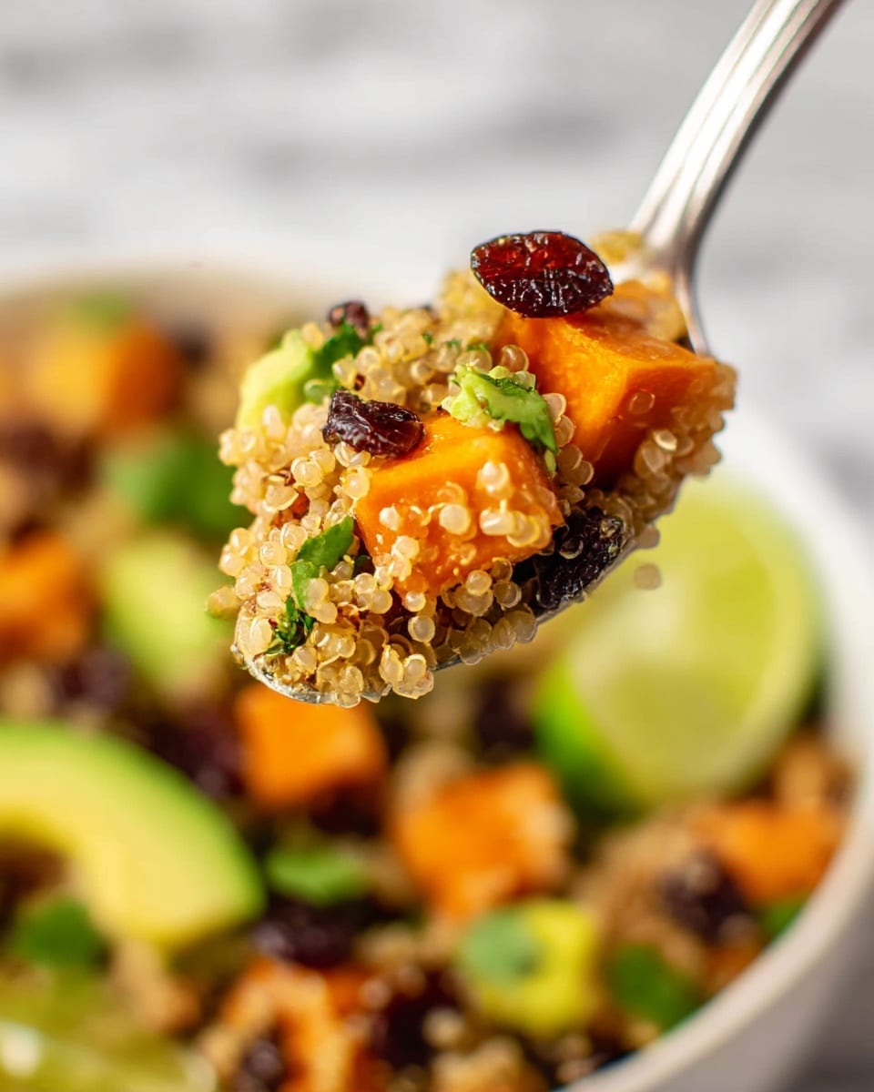 A close-up view of a spoonful of quinoa salad lifted above a white bowl filled with the same salad. The salad has distinct layers of small, round, light beige quinoa grains mixed with bright orange roasted sweet potato cubes, green avocado chunks, dark dried cranberries, and fresh green cilantro leaves. The spoon showcases the quinoa's fluffy texture with visible spirals, along with a piece of dried cranberry and a bit of cilantro on top. In the bowl beneath, the same colorful ingredients appear layered, with lime wedges resting on the side. The whole scene is set on a white marbled surface. photo taken with an iphone --ar 4:5 --v 7