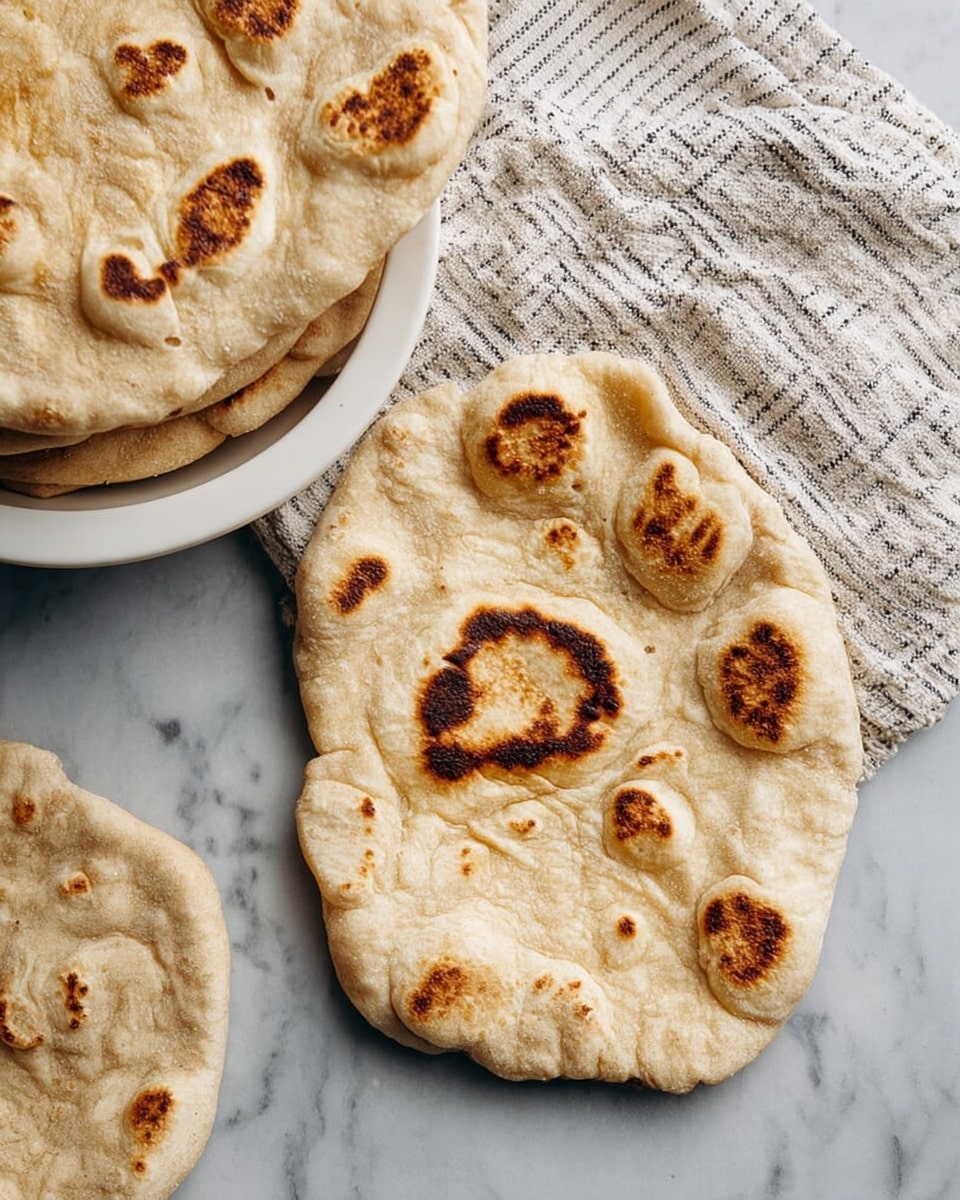 Two pieces of flatbread with uneven, puffy, and bubbly surfaces lie on a white marbled background, each showing several brown charred spots in different shapes and sizes. To the upper left, a white bowl holds more flatbreads stacked partially under a cloth with thin black stripes. The bread has a warm, light brown color with textured grain visible, and the puffed bubbles create an airy, soft look. photo taken with an iphone --ar 4:5 --v 7