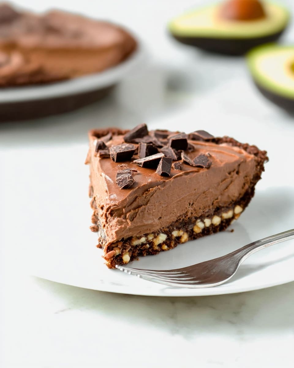A slice of chocolate pie sits on a white plate with a silver fork beside it. The pie has three layers: a dark brown crust made of nuts and chocolate bits at the bottom, a thick smooth milk chocolate filling in the middle, and small chopped chunks of dark chocolate scattered on top. The pie slice is the main focus, placed on a white marbled surface with a blurred full pie and half an avocado in the background. Photo taken with an iphone --ar 4:5 --v 7