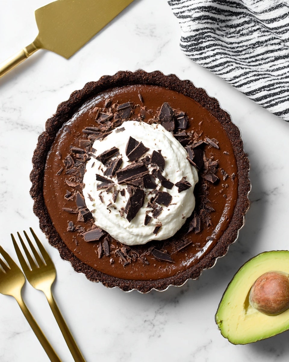 A round chocolate tart with a dark brown crust and a smooth, rich chocolate filling as the first layer. On top of the filling is a dollop of white whipped cream in the center. Dark chocolate shavings of various sizes are scattered over the whipped cream and spread around the tart surface. The tart is on a white marbled surface, with a gold cake server at the top left and two gold forks crossed at the bottom left. On the right side, there is a half-cut avocado showing its light green inside and the pit removed. A black and white striped cloth is partially visible in the top right corner. Photo taken with an iphone --ar 4:5 --v 7