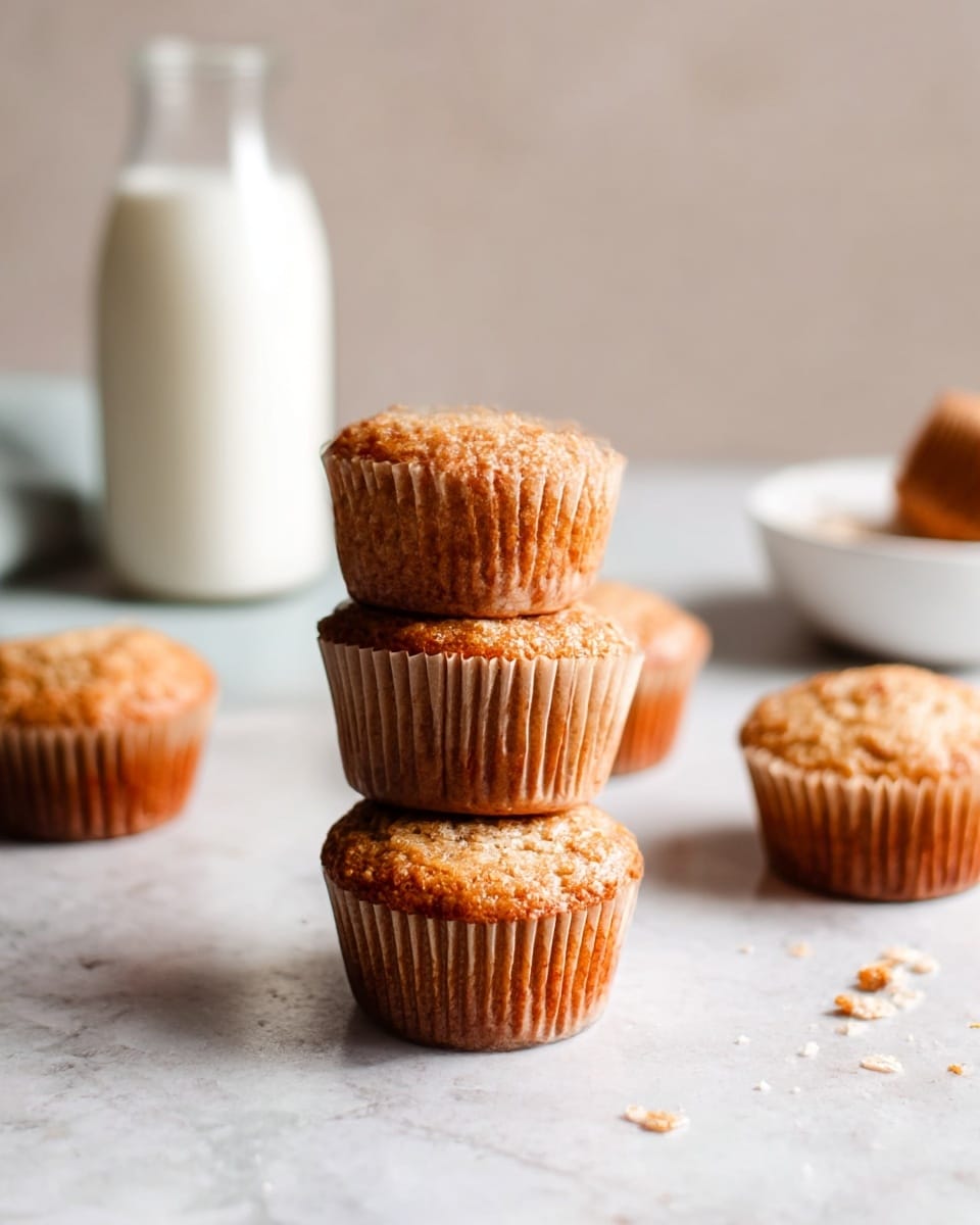 A close-up of six golden-brown muffins with a textured, slightly crisp top and soft paper wrappers, two of which are stacked in the front center while the other four stand alone in the background. The muffins sit on a surface with a white marbled texture scattered with a few small crumbs. In the blurred background, there is a glass bottle filled with white milk and a white bowl partially visible. The colors are warm and natural, with soft lighting giving the muffins a fresh, homemade look. Photo taken with an iphone --ar 4:5 --v 7