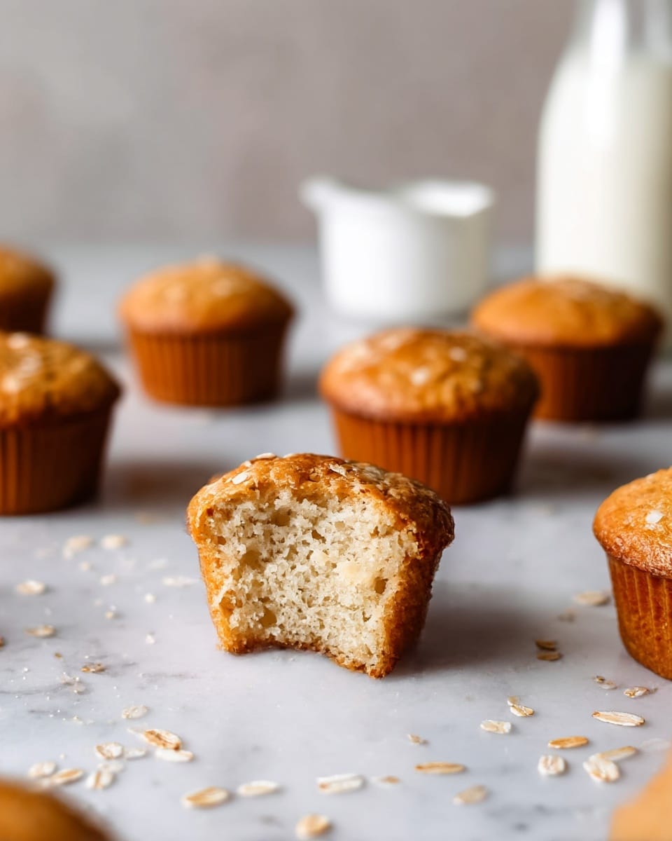 A close-up of a small golden brown muffin with a bite taken out of the front, showing its soft, light, and fluffy pale inside texture; the muffin is set on a white marbled surface scattered with small bits of oats. Around it, there are six whole muffins with a cracked top and a crispy edge, also golden brown, placed in random spots on the white marbled surface. In the background, there is a clear bottle filled with white milk and a small white ramekin, both slightly blurred. The overall mood is warm and cozy with natural light highlighting the muffins' textures. photo taken with an iphone --ar 4:5 --v 7