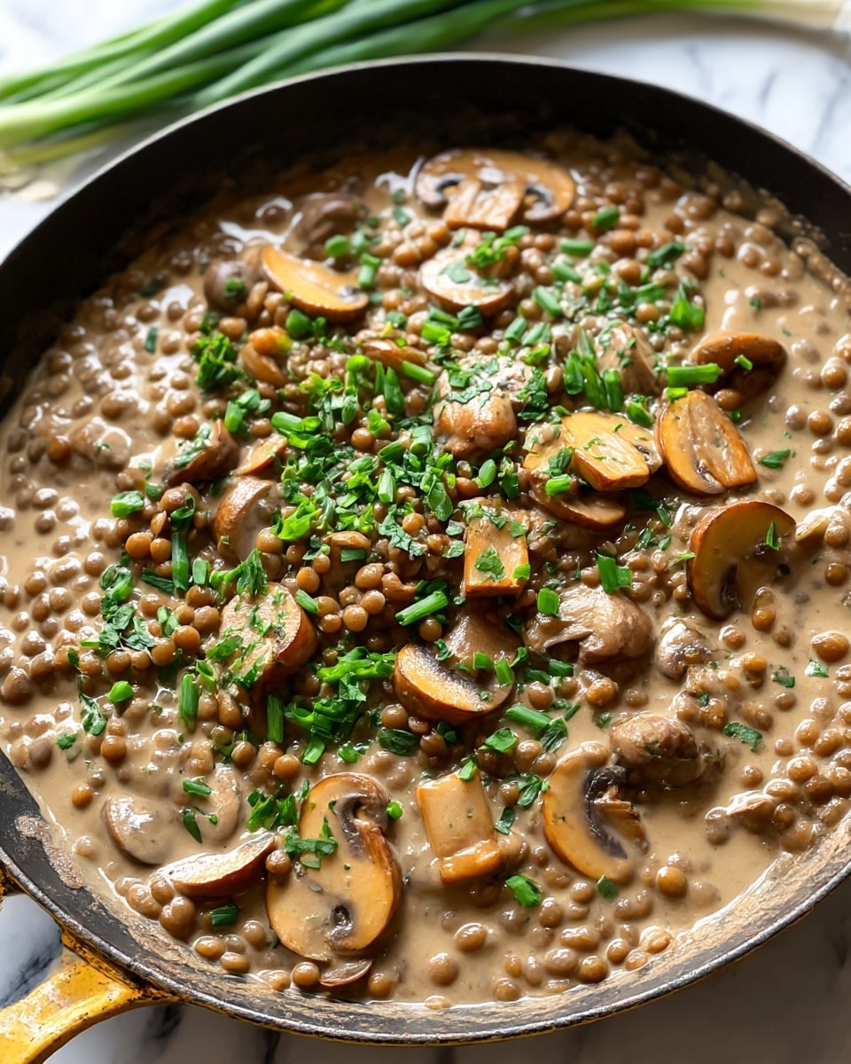 A close-up image of a pan filled with a creamy lentil and mushroom dish, where light brown lentils and sliced golden-brown mushrooms are mixed in a smooth beige sauce, topped with bright green chopped herbs scattered evenly over the top. The pan is dark with worn yellow handles, and there are fresh green onions blurred in the background on a white marbled surface. The texture looks rich and creamy with a mix of soft lentils and tender mushrooms. photo taken with an iphone --ar 4:5 --v 7