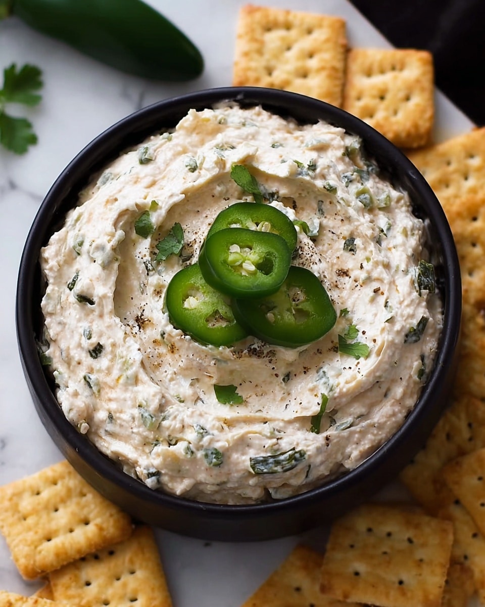 A small black bowl filled with creamy, textured dip speckled with green herb pieces and swirled on the surface, topped with three shiny, sliced jalapeño peppers placed in the center with a few small cilantro leaves underneath. Surrounding the bowl are light tan square crackers with small holes, all set on a white marbled surface. Photo taken with an iphone --ar 4:5 --v 7