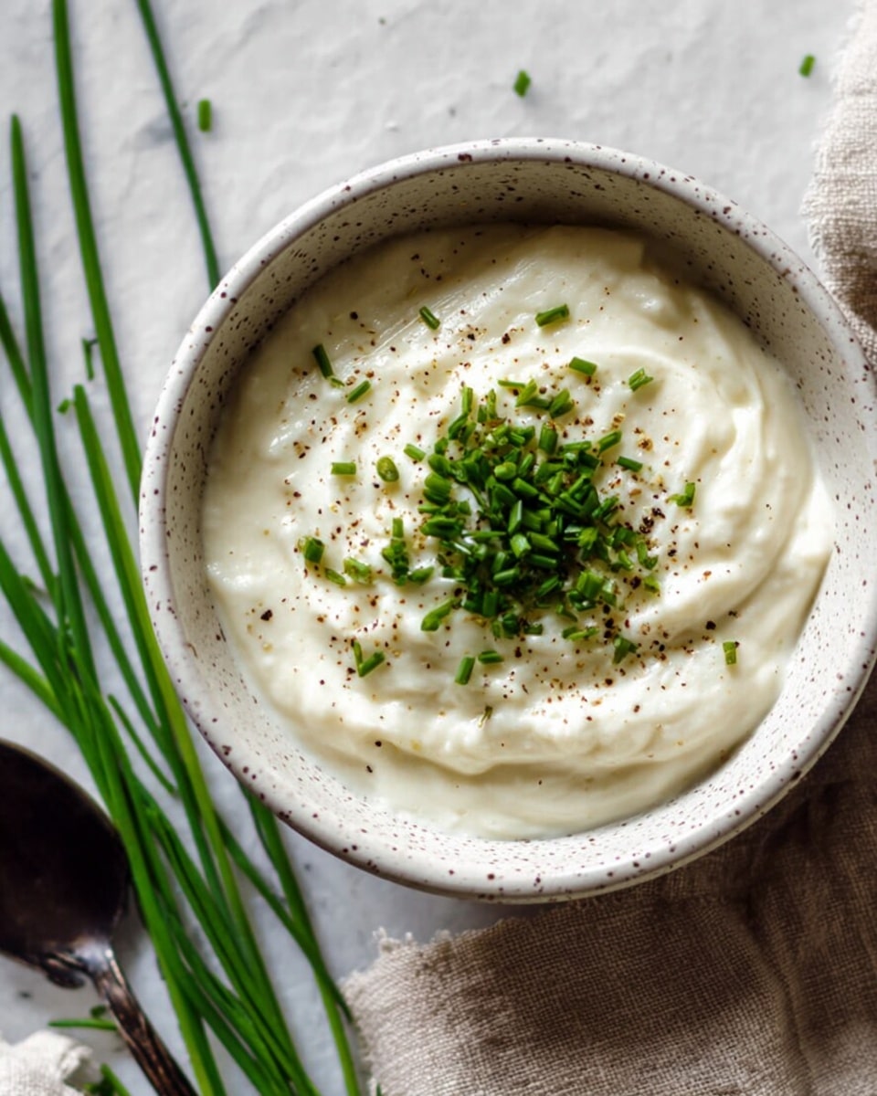 A close-up of a small white speckled bowl filled with thick, creamy white dip that has a smooth, slightly swirled texture. On top, there is a small pile of finely chopped green herbs, likely chives, sprinkled with coarse black pepper, creating a contrast on the creamy surface. The bowl is placed on a white marbled surface next to a beige cloth with green chive stalks resting beside it, and a dark spoon is partially visible on the left side. The scene is softly lit, emphasizing the fresh and simple look of the dip. photo taken with an iphone --ar 4:5 --v 7