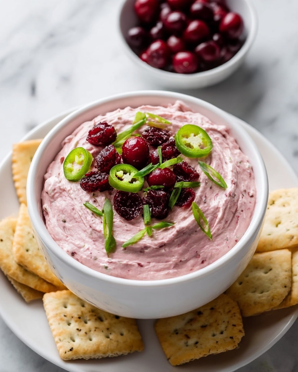 A white bowl filled with two layers of creamy pink dip with a smooth, whipped texture topped with bright red whole cranberries and dark red dried cranberries. On top, scattered bright green chopped scallions and sliced green jalapeños add contrast. The bowl is placed on a white plate holding light golden crackers with visible seasoning. A small white bowl with more dark red cranberries is in the background on a white marbled surface. photo taken with an iphone --ar 4:5 --v 7