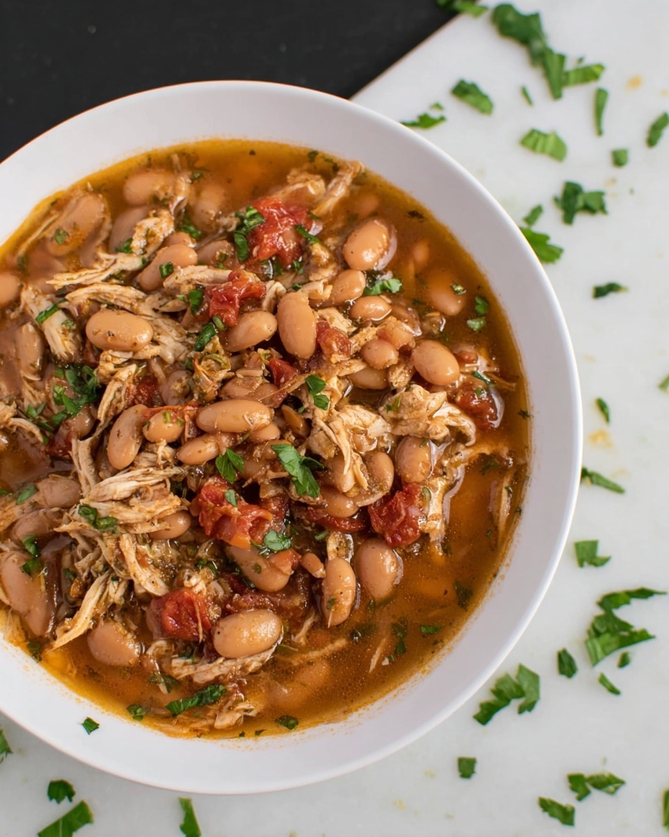A close-up of a white bowl filled with a warm stew made of light brown beans, shredded light beige chicken pieces, and small bits of red tomatoes mixed in a thick, slightly shiny brown broth. Some green herb leaves are scattered on top and around the stew, adding a fresh touch. The bowl sits on a white marbled surface with a few green herb pieces scattered loosely nearby. photo taken with an iphone --ar 4:5 --v 7