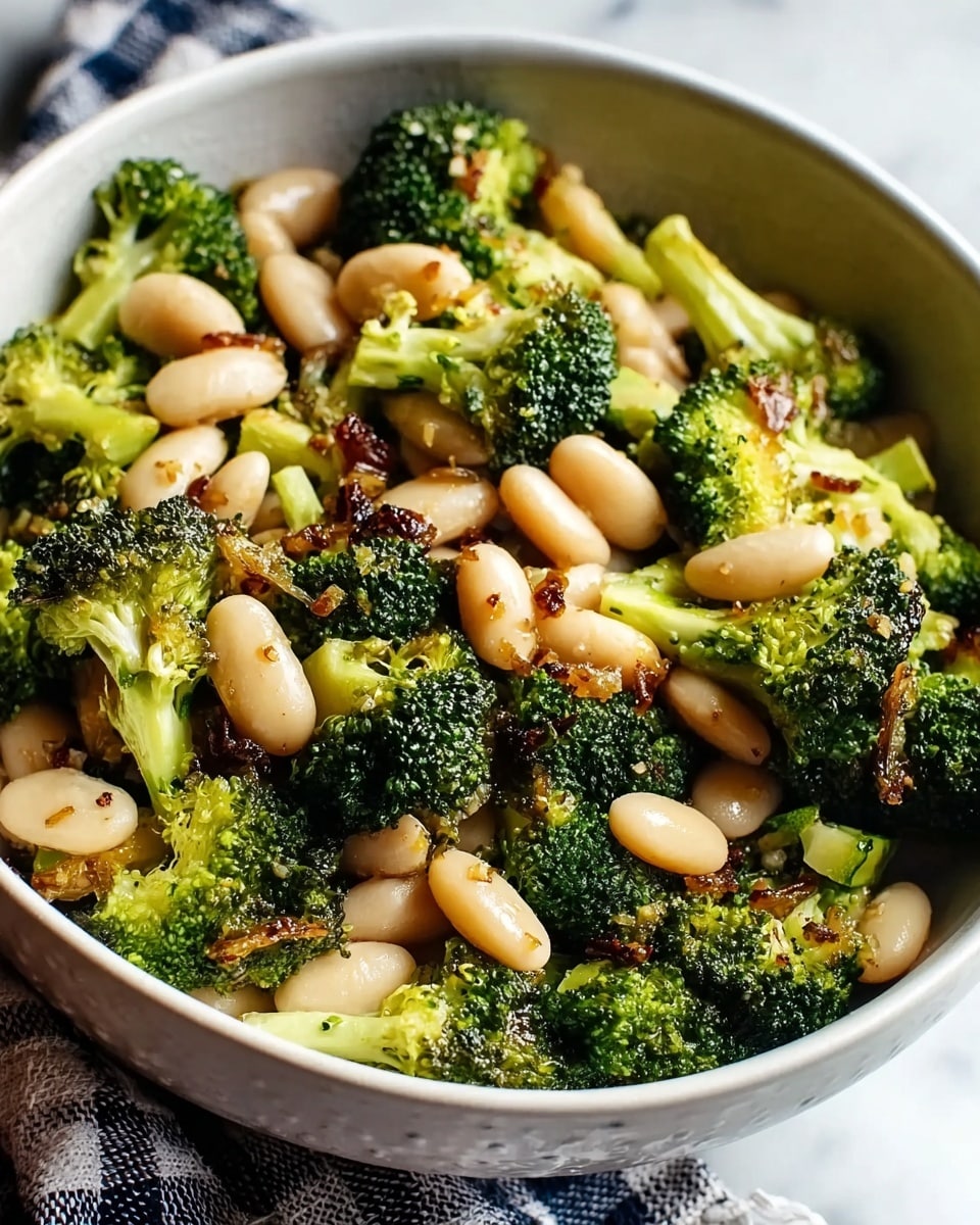 A deep white bowl filled with a mix of roasted broccoli and white beans. The broccoli florets are dark green with some slightly charred edges, showing crisp texture, while the white beans are smooth and soft, scattered evenly throughout. There are small bits of reddish seasoning or spices spread over the vegetables, adding a touch of color. The bowl sits on a white marbled surface, giving a clean and fresh look. photo taken with an iphone --ar 4:5 --v 7