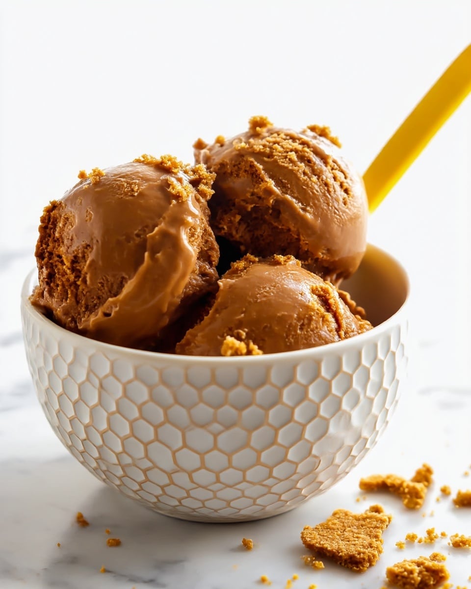 The image shows a white bowl with a honeycomb pattern, filled with three scoops of smooth, rich brown ice cream. The ice cream has a glossy texture with small crumbs of a crumbly biscuit sprinkled on top and around the bowl on a white marbled surface. A yellow spoon is partially visible, sticking out from the side of the bowl. Photo taken with an iphone --ar 4:5 --v 7