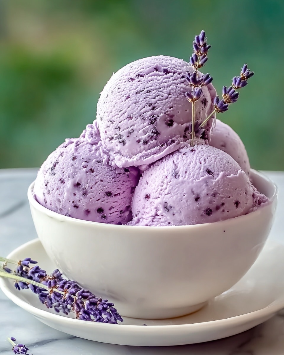 A white bowl filled with four scoops of light purple ice cream with small dark speckles. The scoops are stacked with three at the bottom and one on top, which is decorated with two small sprigs of lavender flowers. The bowl sits on a white plate, and a few lavender flowers rest beside it. The background is a soft green blur, and the surface beneath is a white marbled texture. photo taken with an iphone --ar 4:5 --v 7