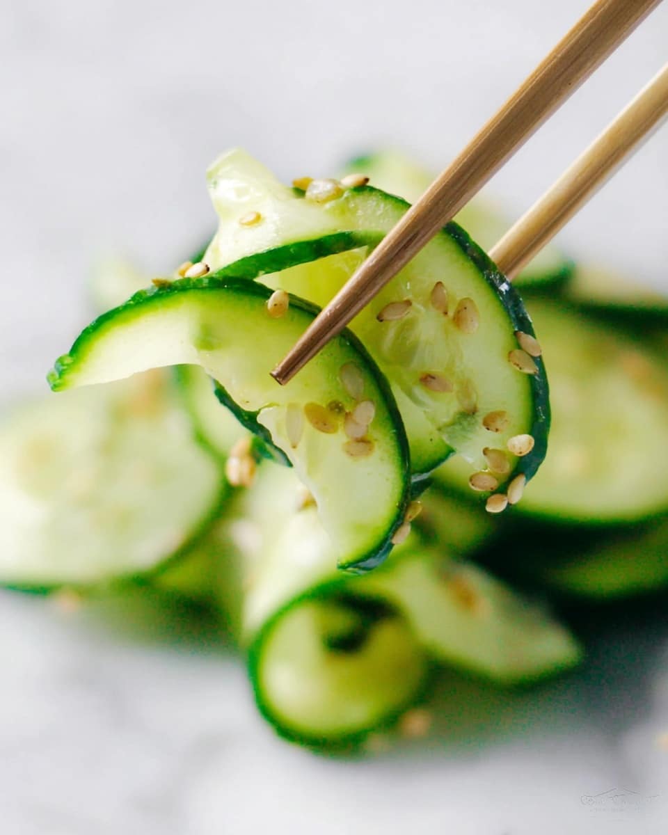 A white bowl filled with many thin slices of cucumber, pale green with dark green edges, layered loosely and topped with small black and light brown sesame seeds scattered on top. A pair of light wooden chopsticks held by a woman's hand is lifting a few curled slices of cucumber from the bowl. In the softly blurred background, a white bowl with whole green cucumbers and orange carrots can be seen, all placed on a white marbled surface. The overall look is fresh and simple. photo taken with an iphone --ar 4:5 --v 7