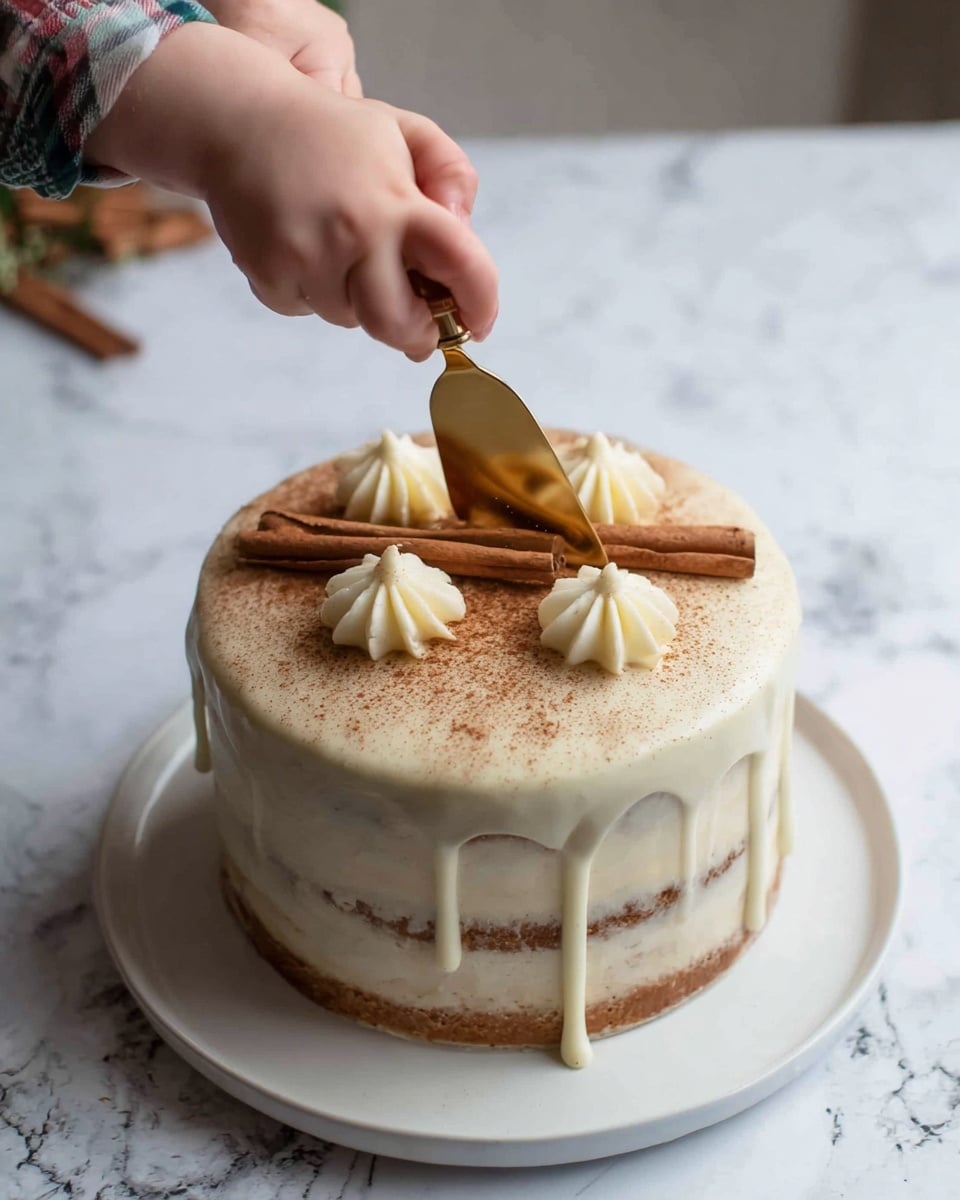 A round cake with three visible layers sits on a white plate on a white marbled surface. The bottom two layers have a light brown color, and the top layer is covered with a smooth creamy white frosting that drips slightly over the edges. The top of the cake is dusted evenly with a light brown powder, likely cinnamon or cocoa, and decorated with four small white frosting swirls arranged in a square. Two cinnamon sticks are placed crossing each other on top of the frosting swirls. A child's small hand and an adult's hand are holding a gold cake server to cut into the cake from the front center. Photo taken with an iphone --ar 4:5 --v 7
