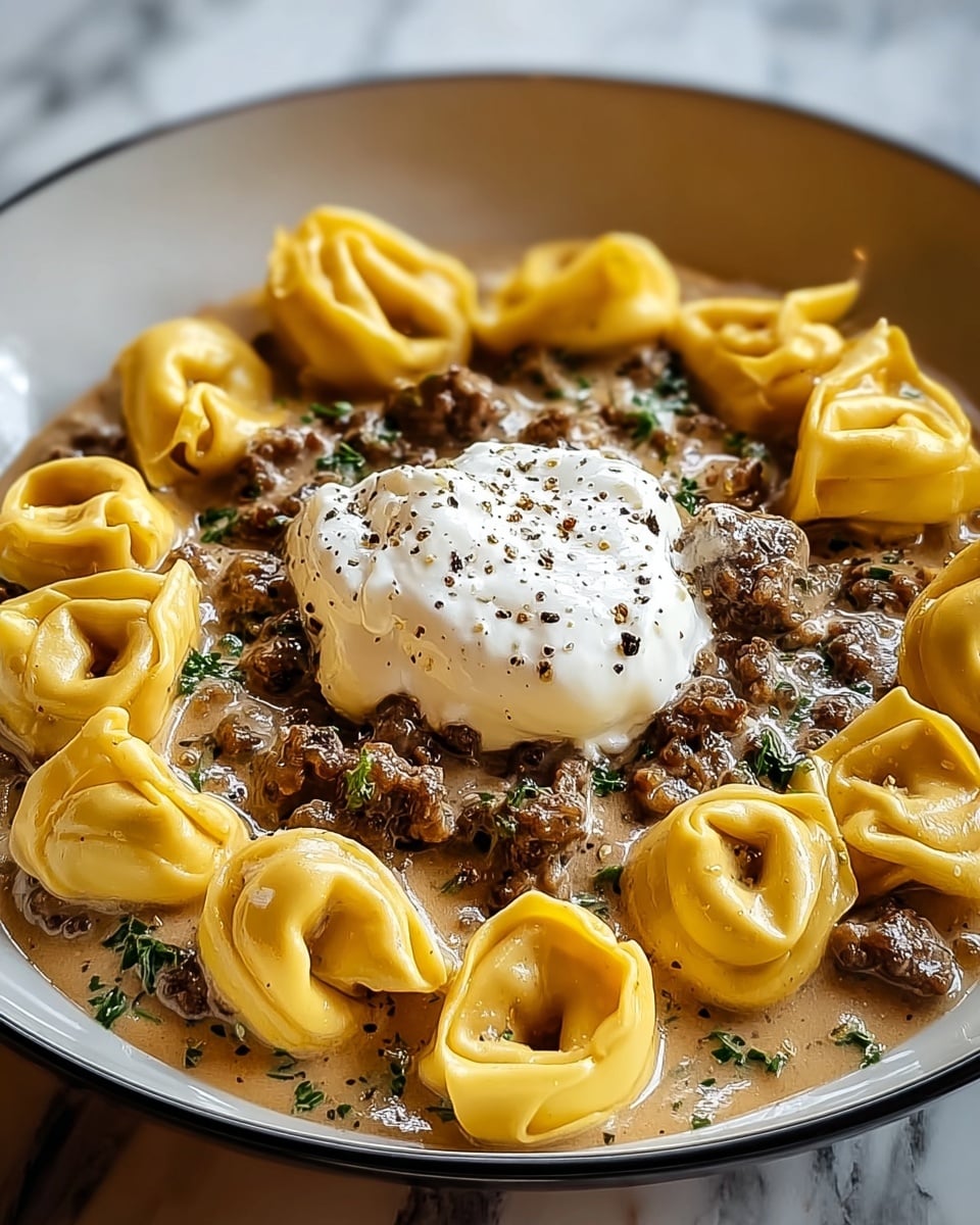 A white plate holds a dish with two main layers and a topping. The bottom layer is a light brown, creamy sauce with small pieces of dark brown cooked meat mixed with green herbs. On top of this sauce, yellow tortellini pasta pieces are arranged evenly, showing their folded, ring shapes with smooth, slightly shiny textures. In the center of the dish, there is a white dollop of soft cream topped with small black pepper specks. The whole meal is served on a white marbled surface. photo taken with an iphone --ar 4:5 --v 7