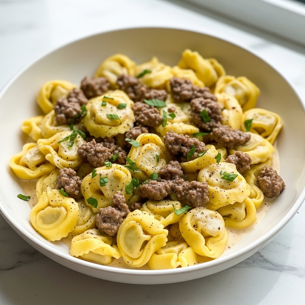 A white bowl filled with yellow tortellini pasta covered in creamy sauce, topped with browned ground meat pieces and small bits of fresh green herbs scattered over the dish. The tortellini appear soft and plump, with smooth, slightly shiny surfaces, while the meat looks crumbly and moist. The sauce pools lightly around the edges, adding a glossy texture, and a light sprinkle of black pepper adds a touch of contrast. The whole dish rests on a white marbled surface near a window with soft natural light. Photo taken with an iphone --ar 4:5 --v 7