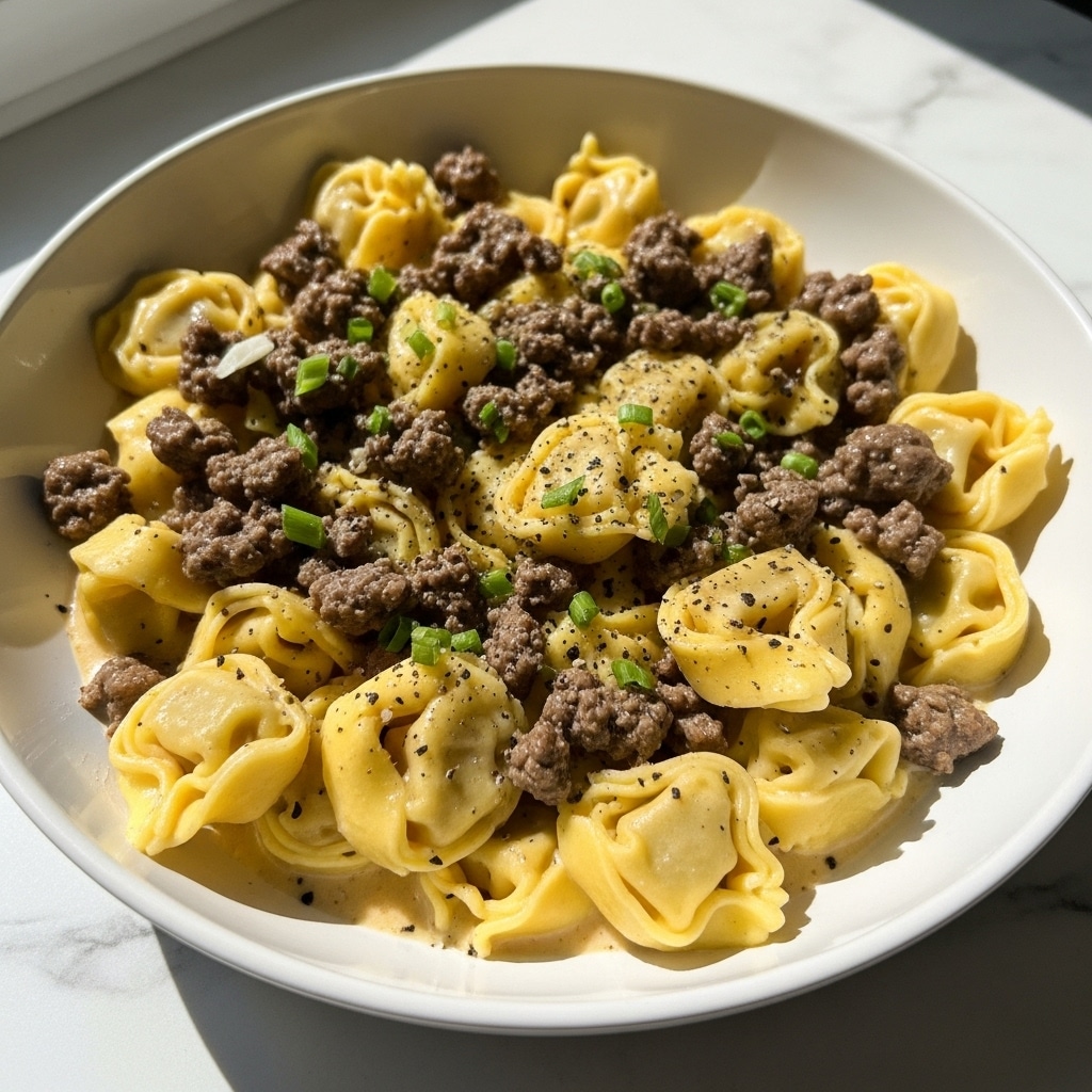 The image shows a white bowl filled with creamy tortellini pasta covered in small pieces of brown ground meat. The tortellini are yellow with a smooth, slightly glossy texture, arranged closely together. The dish is topped with a sprinkling of green chopped herbs and black pepper, adding texture and color contrast. A light, creamy sauce pools slightly at the bottom of the bowl, giving the dish a rich, juicy look. The bowl is placed on a white marbled surface with soft natural light illuminating the scene from a nearby window. photo taken with an iphone --ar 4:5 --v 7