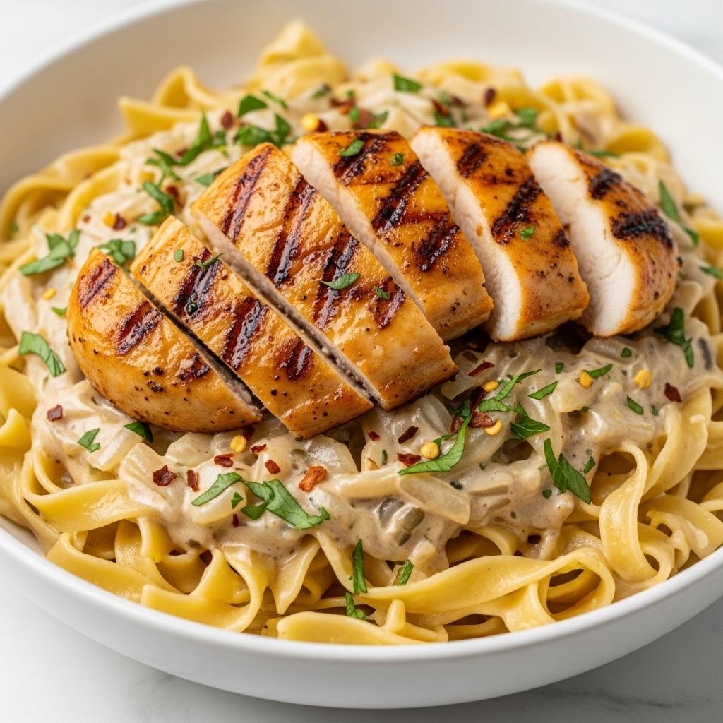 The dish shows a close-up of wide egg noodles covered in a creamy white sauce with herbs and red pepper flakes mixed throughout. On top, there are several pieces of golden-brown grilled chicken breast arranged in a scattered pile, each piece showing a slightly charred texture with a glossy finish. The noodles beneath are soft and slightly curled, coated well in the sauce, with the herbs sprinkled evenly over both the noodles and chicken. The food is served in a white bowl, placed on a white marbled surface. The lighting highlights the glistening sauce and the golden tones of the chicken, giving the dish an inviting look. photo taken with an iphone --ar 4:5 --v 7