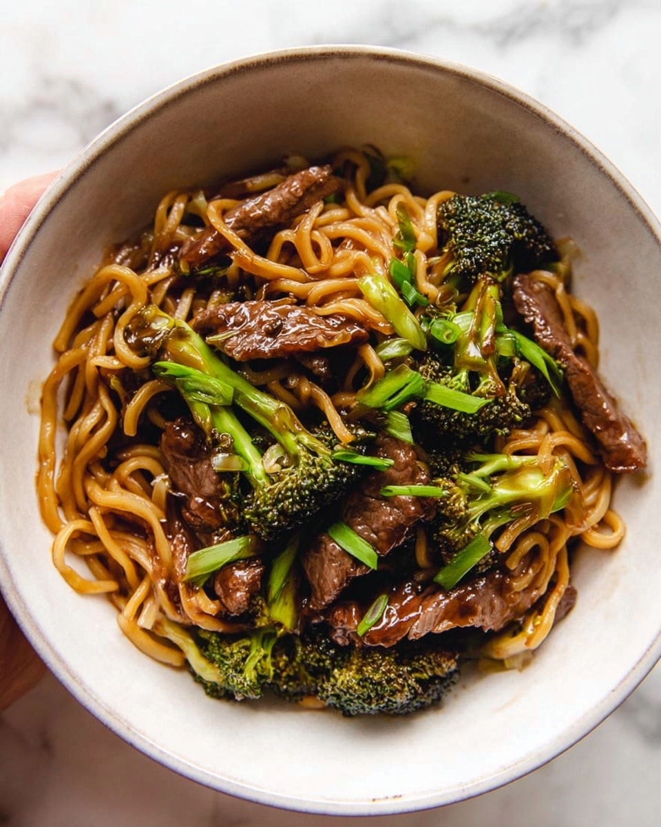 A white bowl filled with three main layers: the bottom layer is thin, light brown cooked noodles tangled around the bowl, the middle layer includes dark brown strips of cooked beef mixed evenly throughout, and the top layer has bright green broccoli florets and sliced green onions scattered on top, all coated in a glossy brown sauce that gives a shiny texture to the noodles and beef. The bowl sits on a white marbled surface with a woman's hand holding the side of the bowl slightly visible. photo taken with an iphone --ar 4:5 --v 7