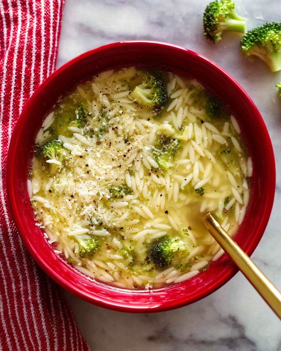 A close-up of a red bowl filled with light yellow broth soup with small white orzo pasta pieces and small bright green broccoli florets mixed in. The soup surface is sprinkled evenly with finely grated white cheese and black pepper flakes. A gold spoon is partially dipped into the soup on the right side, and small broccoli florets are scattered on the white marbled surface near the top right corner. A red and white striped cloth is placed under the bowl on the bottom left side. photo taken with an iphone --ar 4:5 --v 7