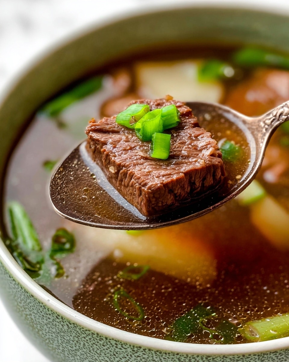 A close-up image of a spoon holding a square piece of cooked beef, brown in color with a slightly textured surface, garnished with two small bright green chopped scallions on top. The spoon is above a bowl of clear brown broth that contains slices of light beige root vegetables and scattered bright green scallions. The bowl is white with a subtle textured, speckled green outer surface, set on a white marbled background. The soup looks warm and fresh with visible tiny oil droplets on the broth surface. photo taken with an iphone --ar 4:5 --v 7