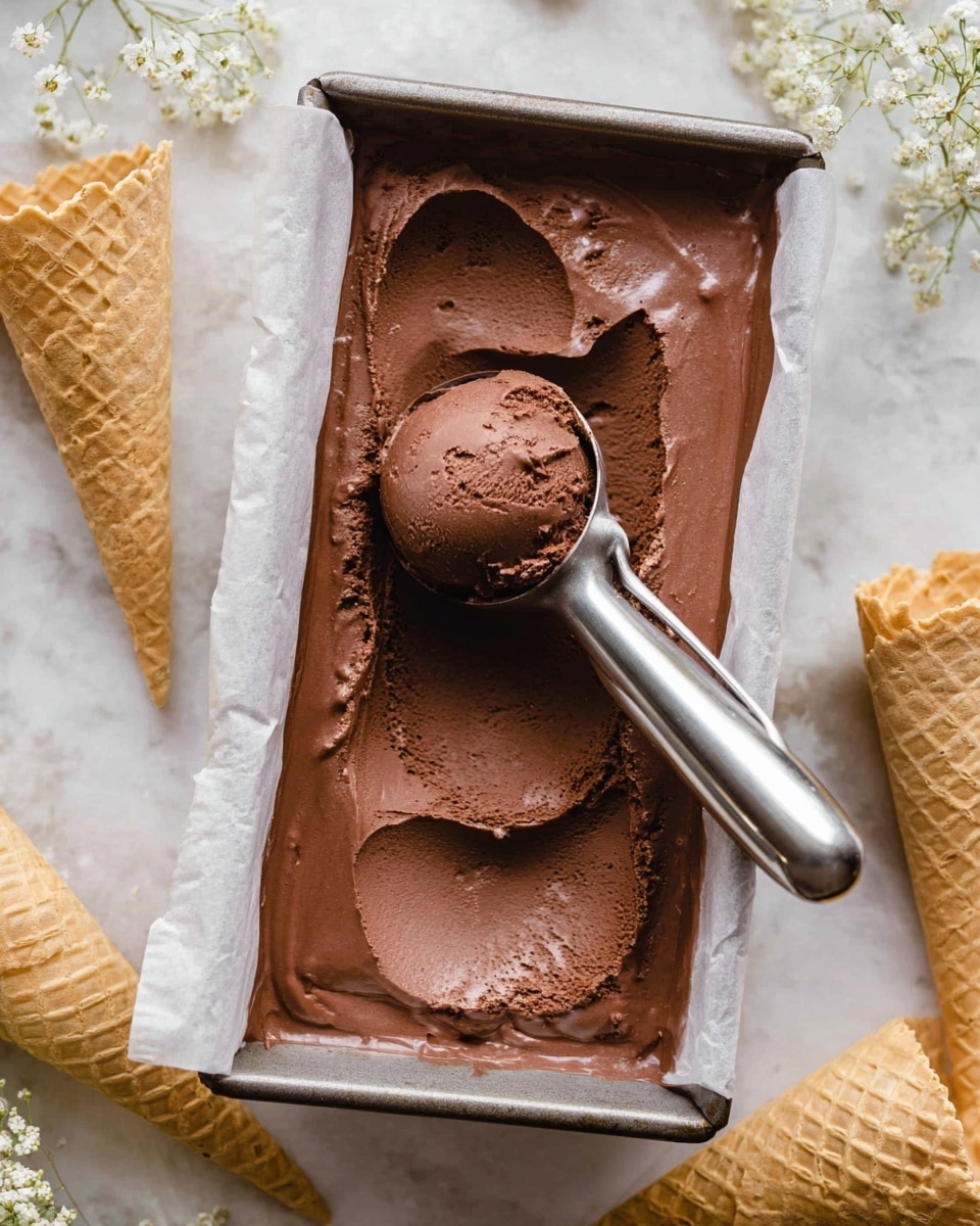 A close-up view of a rectangular metal pan lined with white parchment paper, filled with a rich, smooth, dark brown chocolate ice cream that has a creamy texture. A shiny silver ice cream scoop is lifting a perfect round scoop of the chocolate ice cream from the middle of the pan, showing the smooth, dense inside. The pan is placed on a white marbled surface, surrounded by empty light beige waffle cones and small white flowers as decoration. Photo taken with an iphone --ar 4:5 --v 7