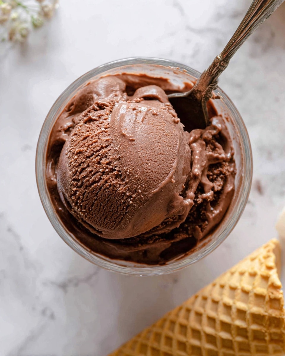 A close-up top view of a single large scoop of rich, creamy chocolate ice cream in a clear glass cup. The ice cream has a dark brown color with a smooth, slightly glossy texture, showing some gentle ridges and soft melting spots on top. A vintage silver spoon is placed inside the cup on the right side, with its handle visible and resting against the edge. The clear glass cup sits on a white marbled surface. On the right side, part of a stack of light golden waffle cones is visible, slightly out of focus. Photo taken with an iphone --ar 4:5 --v 7