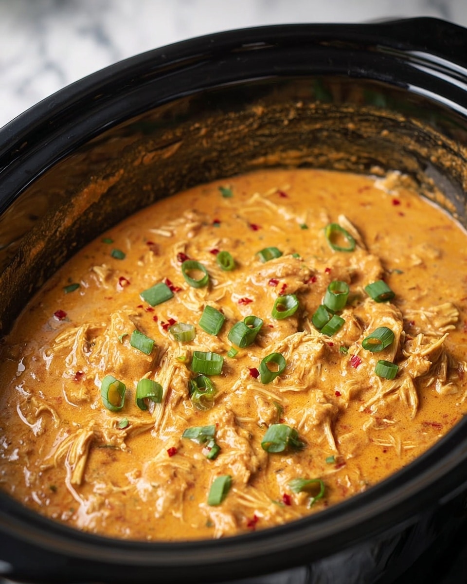 A close-up view of a black slow cooker filled with a thick, creamy orange stew mixed with shredded pieces, likely chicken, throughout. The stew has small red chili flakes and is topped with scattered, chopped bright green onion pieces. The inside edge of the slow cooker shows some splatters of the stew, and the whole dish sits on a white marbled surface. photo taken with an iphone --ar 4:5 --v 7