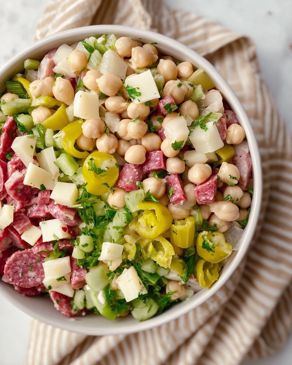 The image shows a close-up of a white bowl filled with a mixed salad. The salad has three main layers: the base is light beige chickpeas and white beans, the middle layer includes bright green celery and chopped parsley, and the top layer features yellow-green slices of pepper rings and small cubes of white cheese. There are also small pieces of pink salami scattered throughout. The bowl is placed on a soft beige and white striped cloth over a white marbled surface. photo taken with an iphone --ar 4:5 --v 7