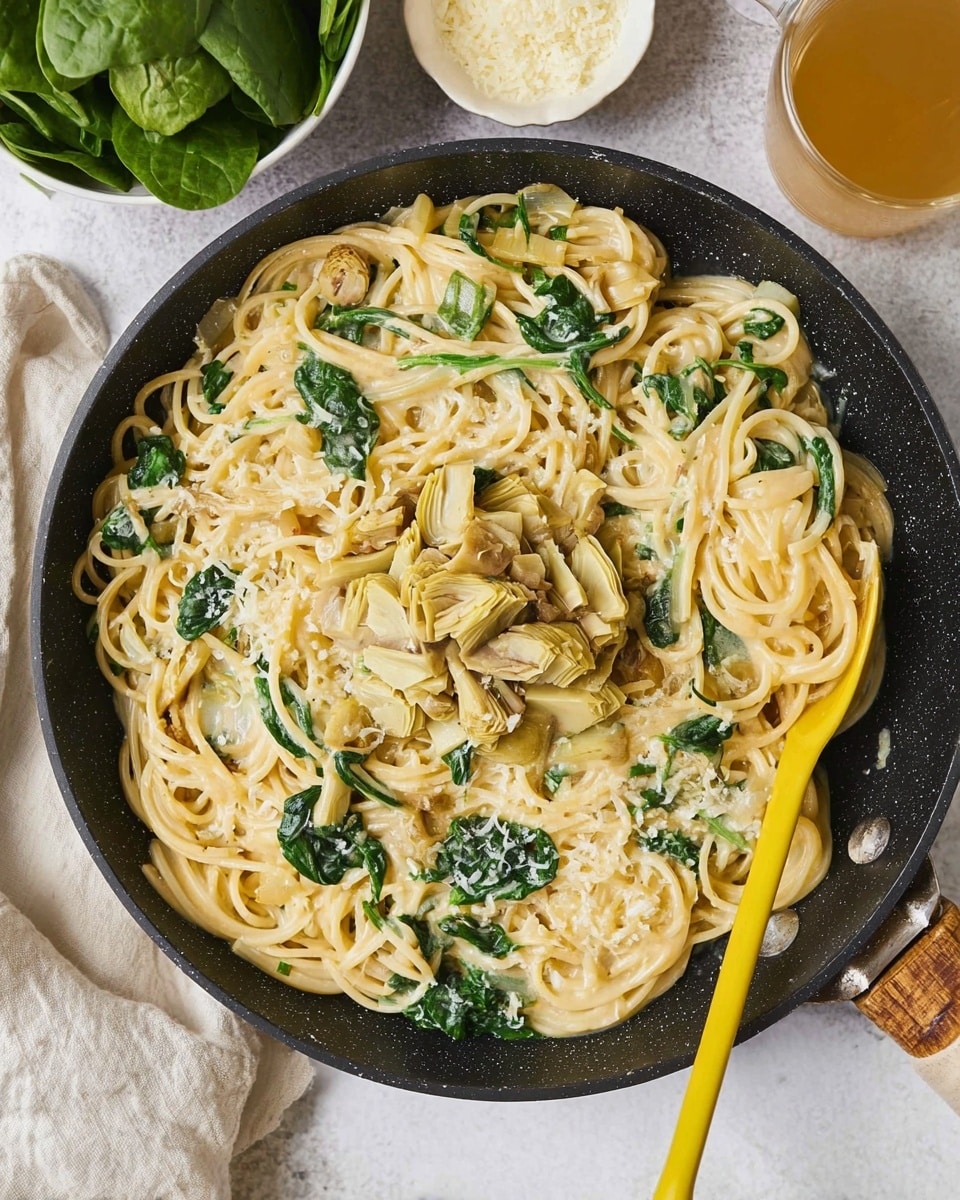 A black shallow pan holds creamy spaghetti pasta mixed with bright green spinach leaves scattered throughout. The pasta is coated in a light, smooth sauce, pale beige in color, with a chunky topping of chopped artichoke hearts placed neatly in the center. A large yellow spoon rests on the right side inside the pan, partially submerged in the pasta. The pan is set on a white marbled surface, with a white bowl of fresh spinach leaves at the top left, a small white bowl of finely grated cheese above the pan, and a glass cup of light-colored tea on the top right. photo taken with an iphone --ar 4:5 --v 7