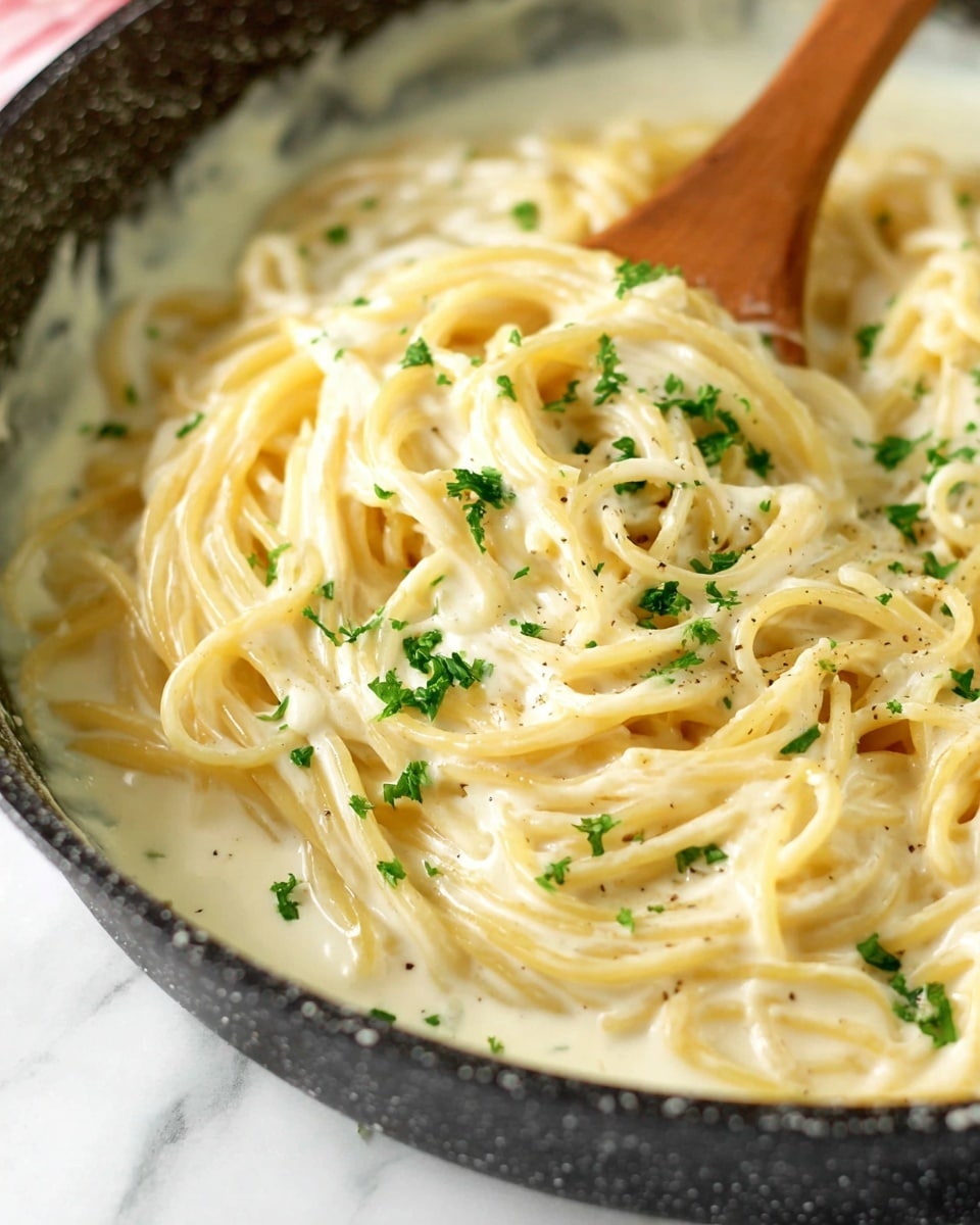 A close-up view of creamy spaghetti pasta in a skillet, showing a thick, smooth white sauce coating the long, slightly yellow noodles. Bright green chopped parsley is sprinkled over the pasta, adding small pops of color. A wooden spoon is stirring the pasta from the right side, mixing the sauce and noodles together. The skillet has a dark textured edge, and the background shows a white marbled texture. photo taken with an iphone --ar 4:5 --v 7