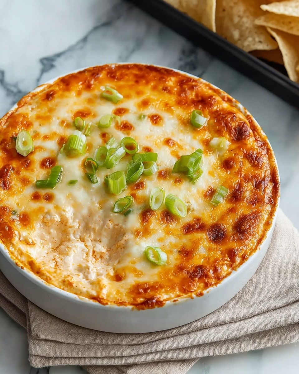 A round white ceramic dish filled with a creamy, cheesy dip that has a golden-brown baked top layer, showing some browned spots. The top layer is melted cheese, slightly bubbly and smooth with small patches of darker browning. Scattered green sliced scallions sit on top, adding a fresh green touch. The dip itself is thick and creamy, with an orange-tinted creamy base visible around the edges. The dish rests on a white marbled surface with a beige cloth napkin beside it. In the blurred background, white tortilla chips can be seen resting on a black tray. photo taken with an iphone --ar 4:5 --v 7