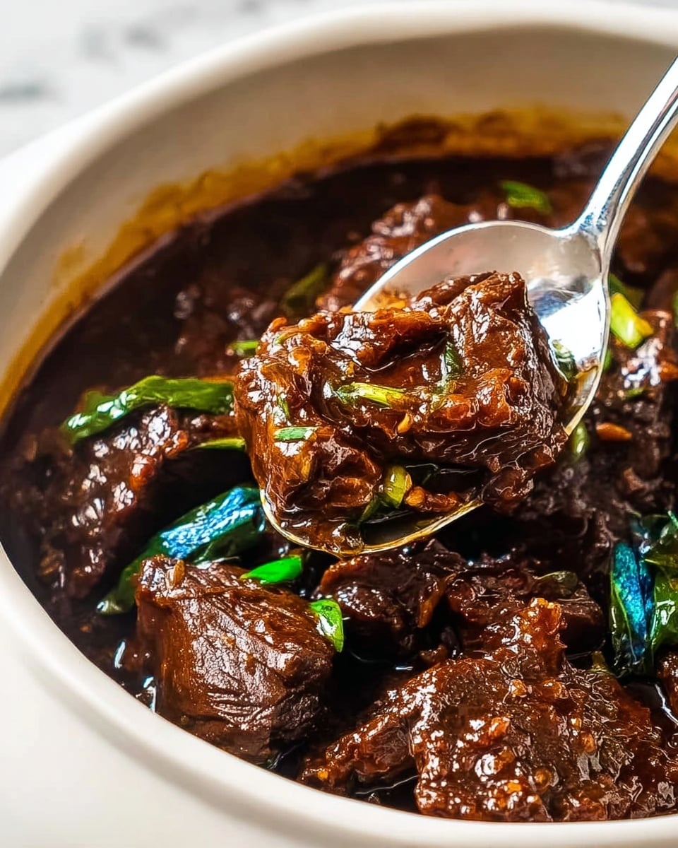 A close-up image shows several pieces of dark brown beef in a thick, glossy sauce with a rich texture inside a white bowl, with a silver spoon lifting one piece above the rest. The beef looks tender and coated in a dark, shiny sauce with some small green vegetable pieces, including leaves with bright blue and green hues, sitting beneath the beef. The white bowl contrasts with the dark food, and the setting has a white marbled texture background. photo taken with an iphone --ar 4:5 --v 7